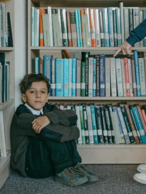 A sad boy sitting on the library floor while being bullied by a peer.