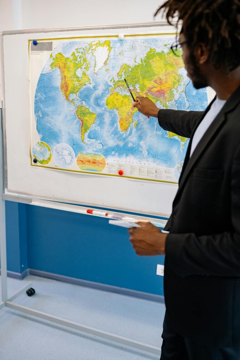 Teacher pointing at world map in classroom setting, emphasizing global geography.